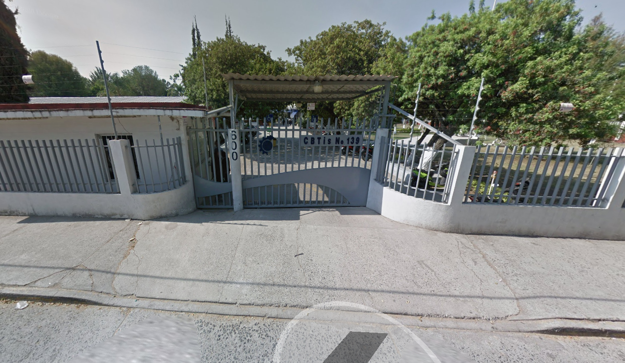 Fotografía en la que se muestra la entrada del plantel, conformada por rejas de color gris que rodean las instalaciones. La reja de en medio está techada y es la que da el acceso al plantel.